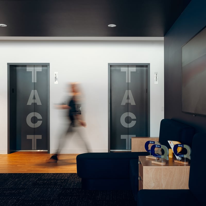 Une personne en mouvement passe devant deux ascenseurs gris portant les lettres T-A-C-T dans un bureau moderne. Au premier plan, un coin salon avec des fauteuils foncés et des trophées sur une table en bois clair.